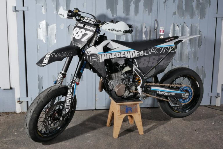 Schwarzes Motorrad mit Independent Racing Dekor vor Metallwand.