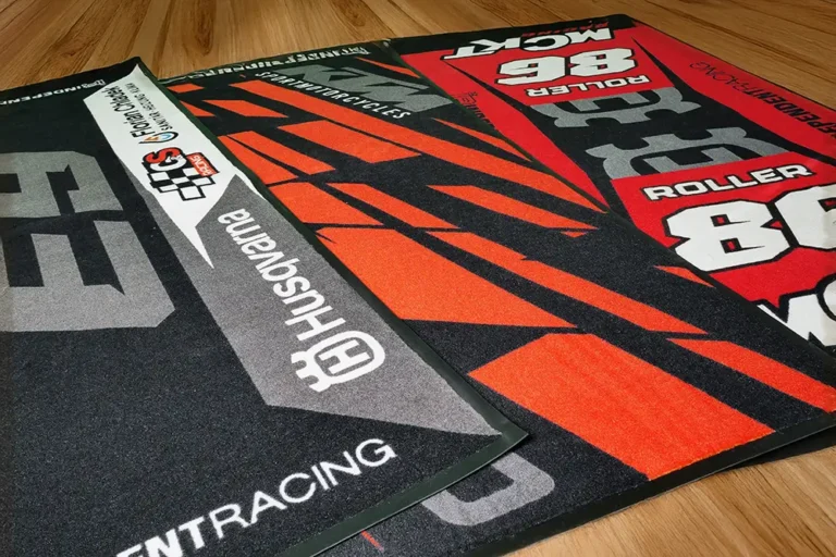 Mehrere bedruckte Motorrad Teppiche in Rot, Schwarz und Grau mit Markenlogos und Startnummern.